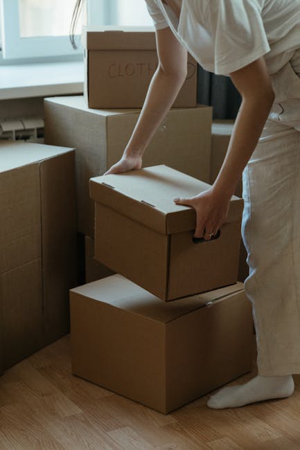 A person wearing light-colored clothing is seen indoors, carefully lifting a cardboard box with a lid, one hand gripping the top and the other supporting the bottom. They are standing on a wooden floo