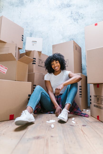 A person wearing light-colored clothing is seen indoors, carefully lifting a cardboard box with a lid, one hand gripping the top and the other supporting the bottom. They are standing on a wooden floo