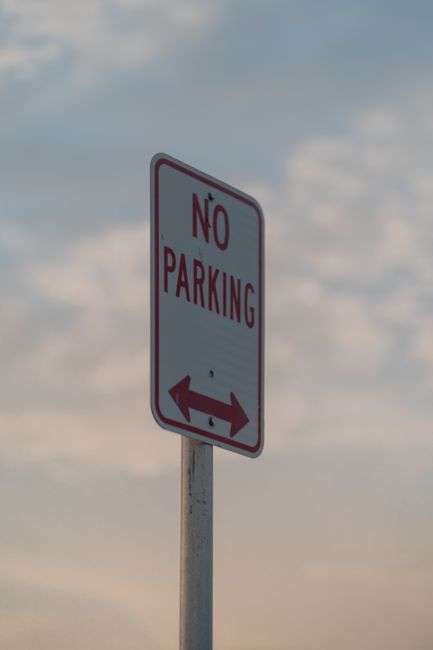 A circular no parking sign with a red border, blue background, and red diagonal line is mounted on a metal pole attached to a brick wall with beige and dark brown stones. The sign casts a shadow on th