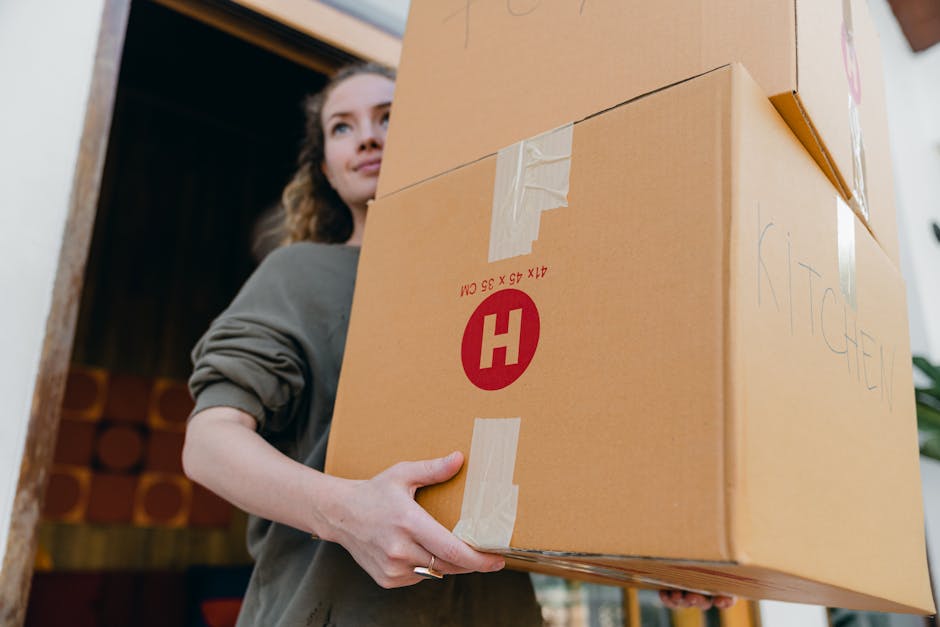 A young woman sits on the floor inside a room surrounded by numerous large cardboard moving boxes, some sealed with red tape and labeled with handling instructions. She appears to be in the process of
